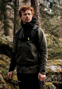 Dark green waterproof jacket with a high collar, black accents, and adjustable cuffs, worn over a black top and paired with dark pants.