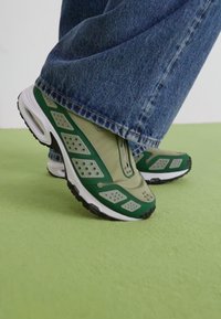 Žalios ir smėlio spalvos sportbačiai su tinklelio detalėmis, traukimo rankena ir storu padu. Tekstūruotos akcentai ir perforacijos šonuose pagerina oro pralaidumą.