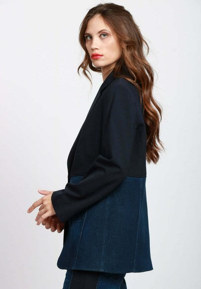 Blazer blu navy scuro con silhouette strutturata e metà inferiore in denim. Tessuto liscio con texture a contrasto e linee pulite.