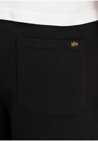 Pantalon de survêtement noir en tissu texturé, avec une poche arrière rectangulaire et un petit logo jaune près de la taille.
