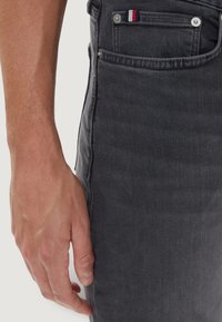 Jeans in denim grigio con un primo piano di una mano appoggiata sulla gamba, caratterizzati da una piccola etichetta tricolore sulla tasca e dettagli classici con bottoni.