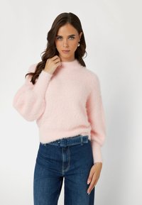 Rosa, flauschiger Pullover mit hohem Kragen und Ballonärmeln, kombiniert mit dunkelblauen, hochgeschnittenen Jeans. Weiches, texturiertes Material.