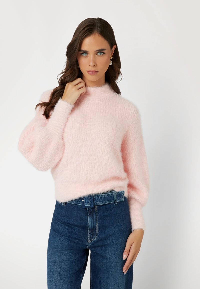 Rosa, flauschiger Pullover mit hohem Kragen und Ballonärmeln, kombiniert mit dunkelblauen, hochgeschnittenen Jeans. Weiches, texturiertes Material.