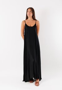 Abito lungo nero realizzato in un tessuto liscio e leggero. Presenta spalline sottili, una scollatura profonda e una silhouette fluida. Indossato con sandali marroni.
