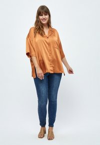 Amber saténová oversized košile s krátkým rukávem, knoflíkovým zapínáním a kapsou na prsou, spárovaná s tmavými džíny skinny a světlými sandály na blokovém podpatku.