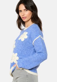 Sininen neulepaita, jossa on valkoisia kukkakoristeita, pyöreä kaula-aukko ja kontrastiväriset helma ja hihansuut. Pehmeä tekstuuri ja rento malli.