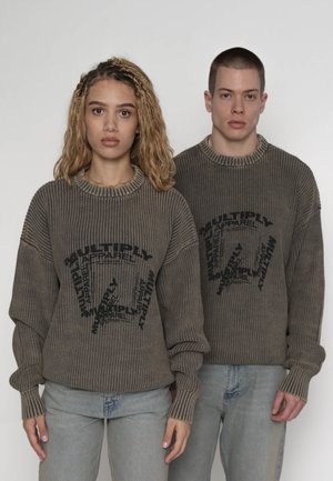 Giovane donna e uomo in piedi fianco a fianco, indossando maglioni grigi oversize coordinati con testo nero e jeans azzurri.