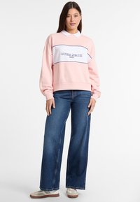Sudadera rosa con panel central blanco, ribete azul y la inscripción "GUESS JEANS 1981". Llevada con jeans de pierna ancha azules y zapatillas blancas.