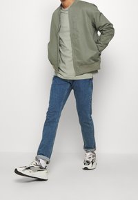 Hellgrüne Bomberjacke, graues T-Shirt, blaue Jeans und mehrfarbige Sneaker. Die Jacke hat gerippte Bündchen, ein minimalistisches Design und einen Reißverschluss.