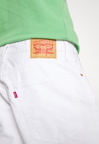Jean blanc Levi's avec une poche arrière visible et un patch en cuir de la marque, porté avec une chemise verte rentrée.