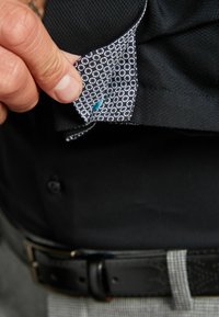 Schwarzes Hemd mit einem gemusterten Innenkragen, der einen blauen Akzent aufweist. Zeigt ein strukturiertes Gewebe und einen sichtbaren Knopf darunter.