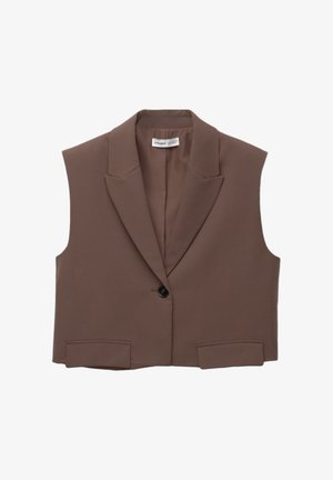 Vrouw draagt een mouwloos, kort bruin vest, wijde bruine broek en bruine sandalen met open tenen, staand tegen een gele muur.