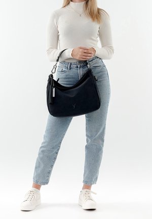 Personne en col roulé blanc et jeans bleu clair tenant un sac à bandoulière noir avec pompon, debout sur fond blanc.