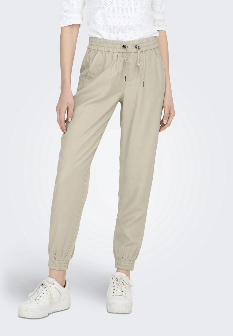 Femme portant un pantalon de jogging beige à cordon, un top en dentelle blanc et des baskets compensées blanches, debout avec un genou légèrement plié.