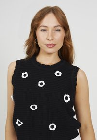 Top senza maniche nero in tessuto texture, con applicazioni di fiori bianchi. Collo con bordi ondulati e vestibilità ampia. Design minimalista.