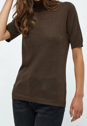 Femme portant un pull à col roulé marron ajusté à manches courtes et un jean bleu foncé, posant devant un fond clair uni.