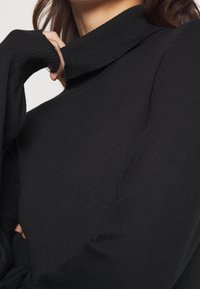 Femme portant un pull à col roulé côtelé noir, une main tirant doucement le col vers le haut près de son cou.