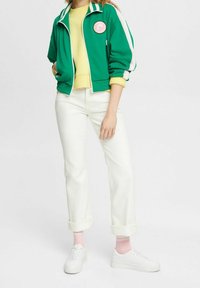 Grön zip-up jacka med vita ränder, gul topp, vita jeans och rosa strumpor. Vita sneakers fulländar outfiten.