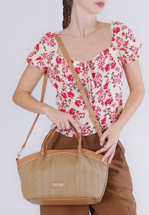 Mujer con blusa floral y pantalones marrones sosteniendo un bolso tejido color canela con correas de cuero sobre el hombro y en la mano.