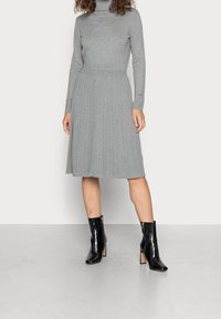 Robe encolure montant grise avec un corsage ajusté, jupes plissées et texture côtelée ; associée à des bottines noires brillantes.
