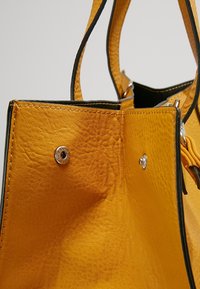 Gul læderhåndtaske med en struktureret overflade og kontrasterende sorte kanter. Har sølvfarvet metalhardware og synlige syninger.