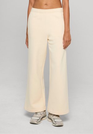 Personne portant un pantalon crème taille haute à jambes larges et des baskets blanches, debout devant un fond uni gris clair.