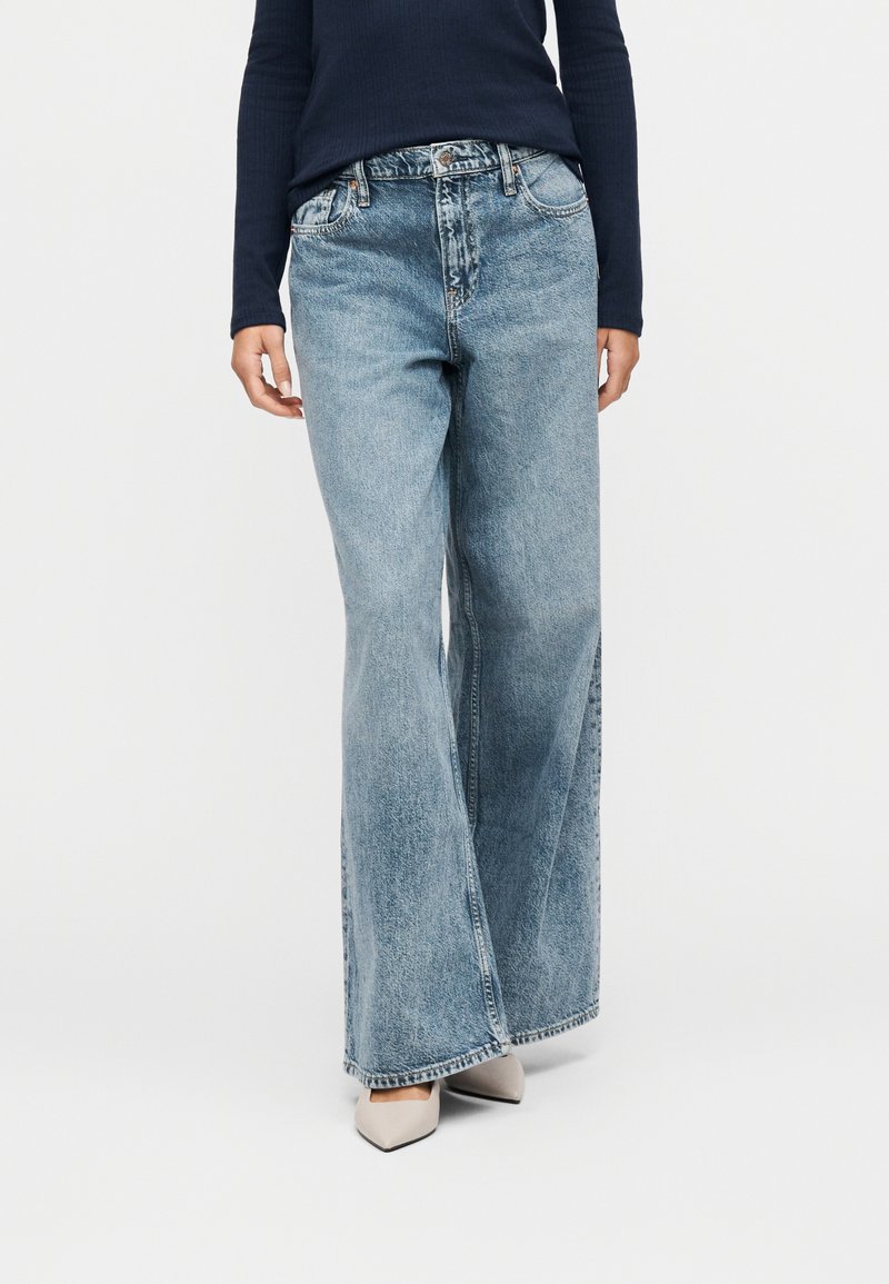 Personne portant un jean taille haute bleu clair et à jambes larges, un haut à manches longues foncé, et des chaussures beiges à bouts pointus sur fond blanc.