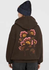 Bruine oversized hoodie met een kleurrijke bloemenprint op de achterkant, met roze, gouden en groene details. Het materiaal lijkt zacht en gestructureerd.