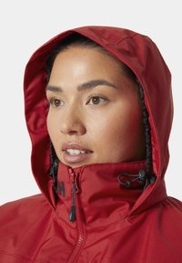 Czerwona wodoodporna kurtka z wysokim kołnierzem i regulowanym kapturem. Posiada czarne zamki i akcenty, z gładką, wytrzymałą teksturą tkaniny.