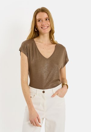 Mujer sonriente con cabello largo y rubio, lleva una blusa metálica marrón con escote en V, pantalones blancos de talle alto, pendientes de aro dorados y una pulsera dorada.