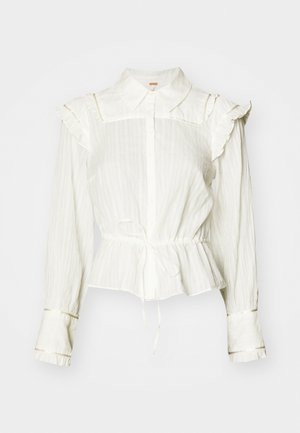 Bílá blůza s dlouhým rukávem, řasenými rameny, knoflíkovým zapínáním a peplum pasem s detailem na zavazování. Materiál je texturovaný a mírně pomačkaný.