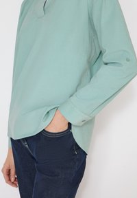 Personne portant une chemise à manches longues vert clair, une main dans la poche d'un jean bleu foncé, sur fond blanc.