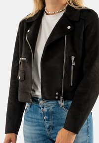Chaqueta de ante negra corta con herrajes plateados, incluyendo cremalleras metálicas y botones a presión, combinada con jeans de mezclilla azul claro.