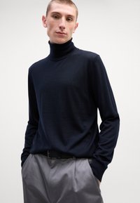 Suéter de gola alta azul-marinho em material de malha suave, com mangas longas e silhueta ajustada, sobre calças plissadas cinzas.
