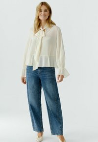 Smiling woman wearing a sheer cream blouse with a neck tie, wide bell sleeves, blue straight-leg jeans, and white pointed heels standing upright.