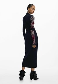 Pikk mereväe sinine kleit, millel on krohvitud korsett ja pikad varrukad, millel on tekstureeritud punase ja roosa mustriga varrukad, kombineeritud mustade kontsadega saapadega.