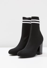 Bottines en tricot noir à talon carré haut. Elles présentent une texture côtelée et un accent rayé blanc en haut.