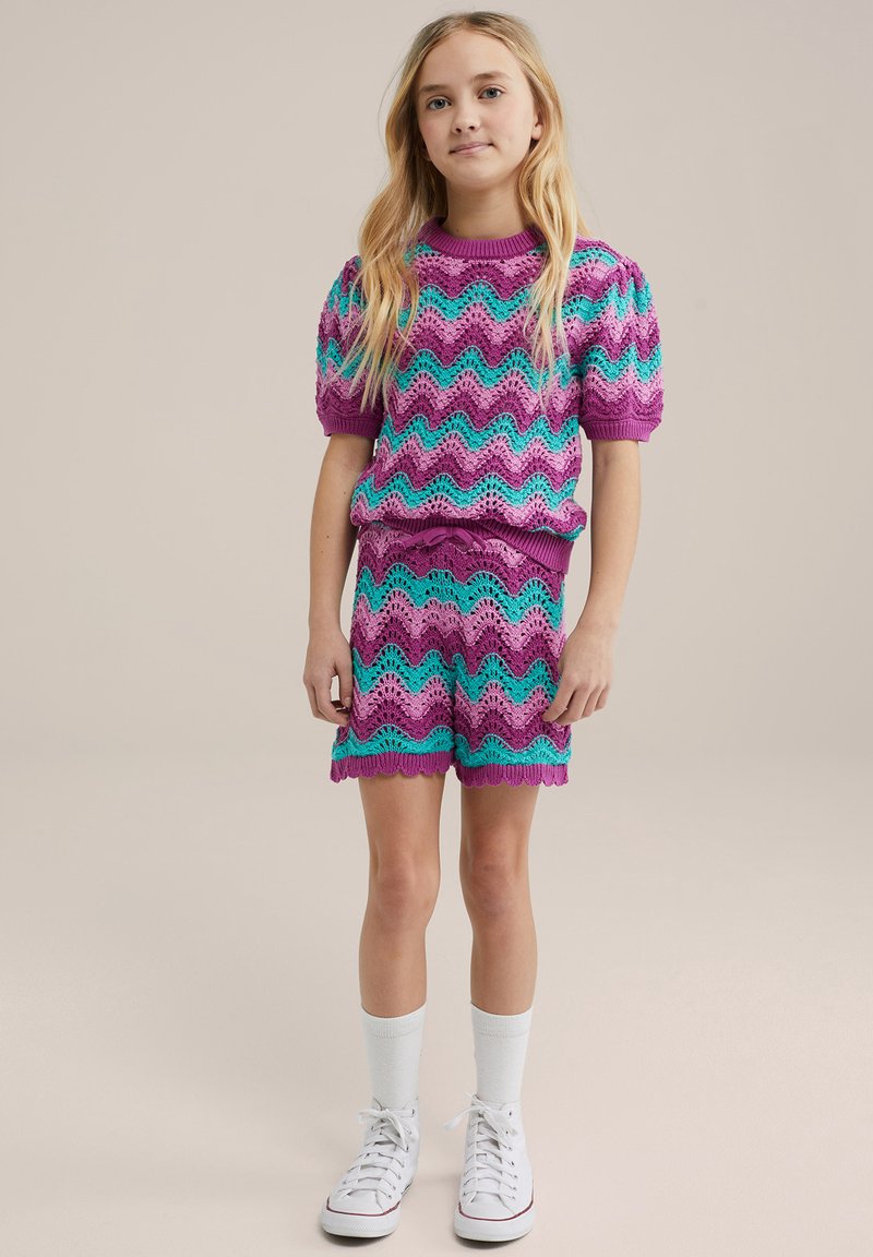 Pleteni dvodelni komplet s valovitim vzorcem v roza, vijolični in turkizni barvi, s kratkimi rokavi in ujemajočimi se hlačami. Bele superge in nogavice.