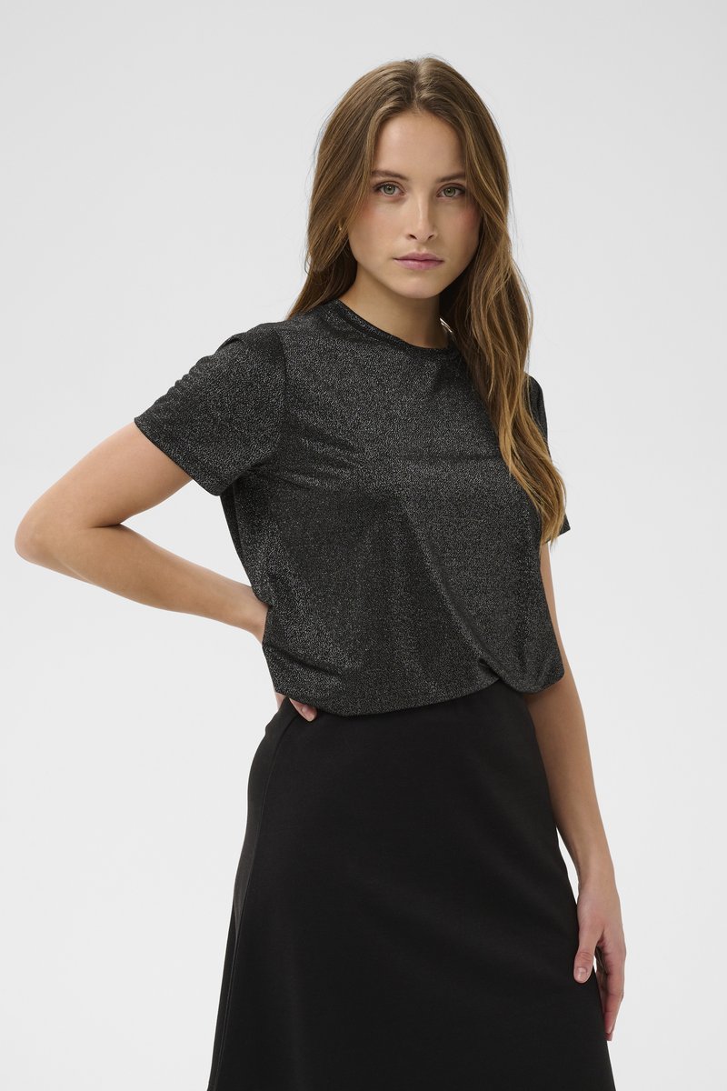 Jauna moteris su ilgais plaukais dėvi trumpomis rankovėmis juodą metalinį viršutinį drabužį ir juodą sijoną, pozuoja viena ranka ant klubo.