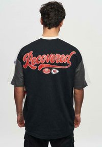 Sort og grå oversized t-shirt med rød "Recovery" tekst på bagsiden, med fodboldgrafik detaljer og rund hals design.