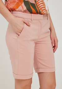 Gaiši rozā pielāgoti šorti, izgatavoti no gludas auduma, ar sānu kabatām un uzrullētām malām, nodrošinot tīru apdari.