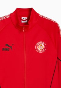 Rød jakke med lynlås har Puma-logo, Girona FC-emblem og "KING" tekst i sort. Dekorative mønstre på ærmerne komplementerer designet.