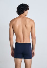 Mørkeblå boksershorts med en tett passform, myk stoff og en midjebånd med logo, sett fra bak. Hud synlig over shortsen.