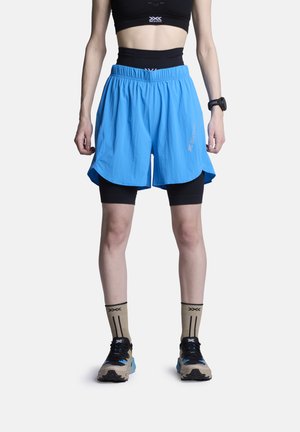 Athlète portant un soutien-gorge de sport noir, un short de compression noir sous un short ample bleu vif, des chaussettes beige et des chaussures de course noires, debout.