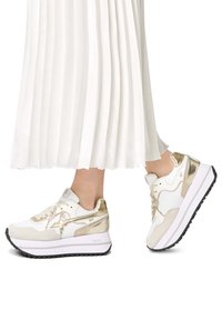 Vita sneakers med guld accenter, med en texturerad ovandel, lagerdesign och en tjock vit sula med ett svart mönster.