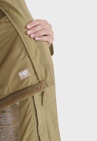 Parka vert olive avec une doublure en shearling marron douce. Comprend une étiquette en tissu, des poches zippées et une texture extérieure lisse.