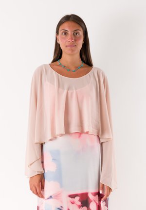 Donna con lunghi capelli castani che indossa un top fluente rosa pallido, una gonna floreale pastello e una collana di perline turchese, in piedi davanti a uno sfondo bianco.