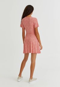 Femme aux longs cheveux bouclés portant une courte robe rouge à motif floral blanc et des baskets beiges, debout de dos sur un fond blanc.