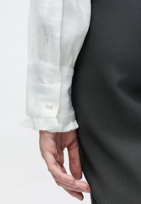 Balta krekls ar volānu aprocēm un divām caurspīdīgām pogām. Roka atbalstīta uz gludas, tumši pelēkas svārku auduma.