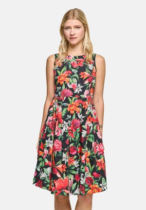 Woman standing, wearing a sleeveless, knee-length dress with a bright, multicolored floral pattern on a black background.